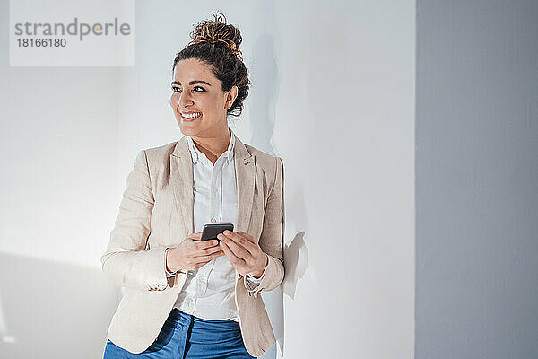 Lächelnde Geschäftsfrau mit Smartphone lehnt an der Wand