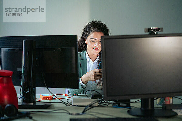 Lächelnde Geschäftsfrau nutzt Mobiltelefon am Desktop-PC am Arbeitsplatz