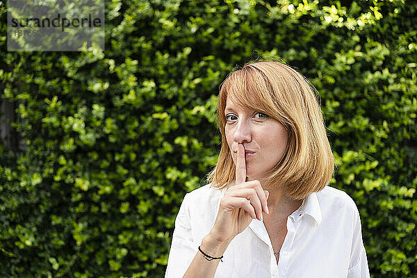 Frau mit Finger auf den Lippen steht vor einer grünen Hecke