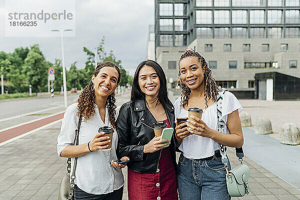 Lächelnde  gemischtrassige Freunde mit wiederverwendbaren Kaffeetassen stehen auf dem Fußweg