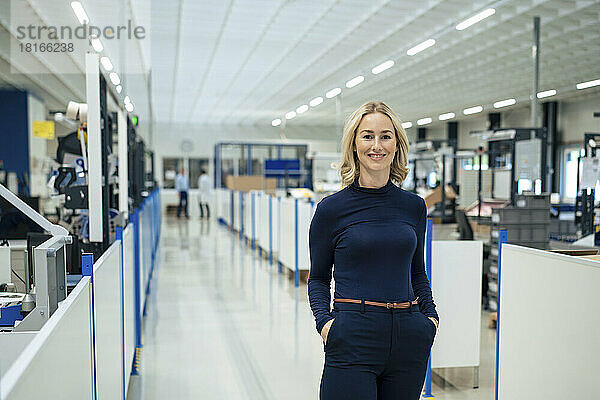 Lächelnde blonde Geschäftsfrau mit Händen in den Taschen in der Fabrik