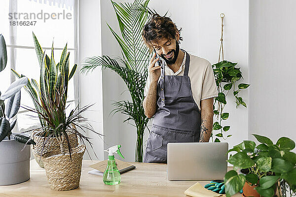 Glücklicher Mann  der im Pflanzenladen mit dem Handy und dem Laptop spricht