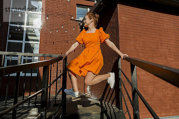 Lächelnde Frau in orangefarbenem Kleid balanciert auf Treppengeländer