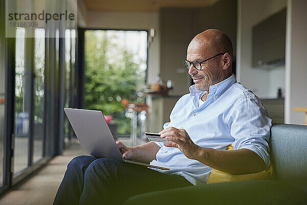 Lächelnder älterer Mann  der zu Hause mit Kreditkarte am Laptop bezahlt