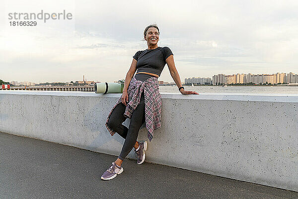 Happy mature woman with exercise mat leaning on wall
