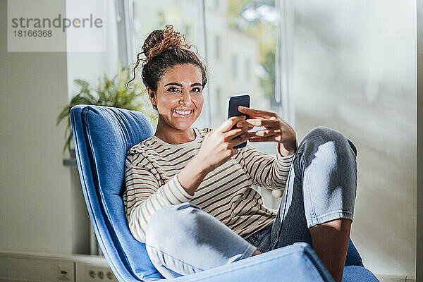 Lächelnde Frau hält ihr Mobiltelefon zu Hause auf einem Stuhl