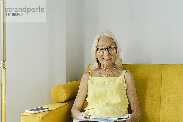 Glückliche ältere Frau  die zu Hause auf dem gelben Sofa sitzt