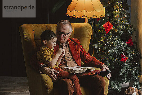 Enkel sitzt auf Großvaters Schoß und liest zu Hause ein Buch