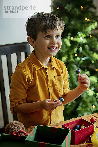 Fröhlicher Junge mit Weihnachtskugeln und Geschenken am Tisch