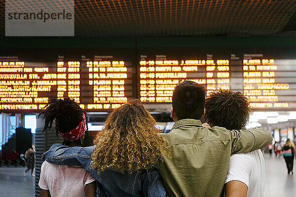 Multiracial friends checking time on departure board at railroad station