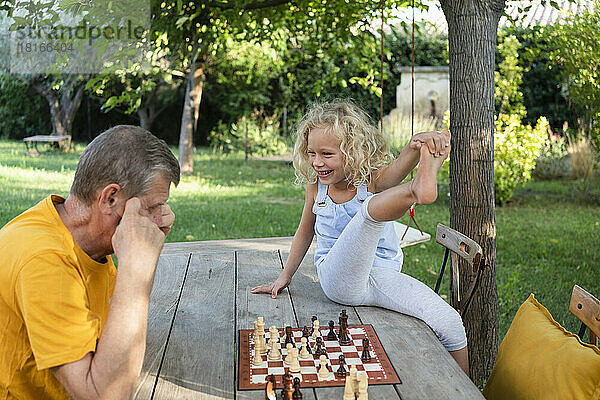 Glückliches blondes Mädchen schaut Großvater beim Schachspielen auf dem Tisch im Garten an