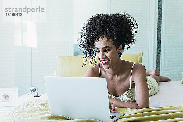 Happy woman with curly hair lying on bed using laptop at home