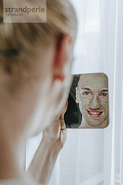 Happy young make-up artist with facial mask looking at mirror