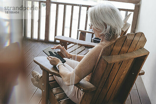Reife Frau sitzt auf einem Stuhl und nutzt einen Tablet-PC auf der heimischen Terrasse