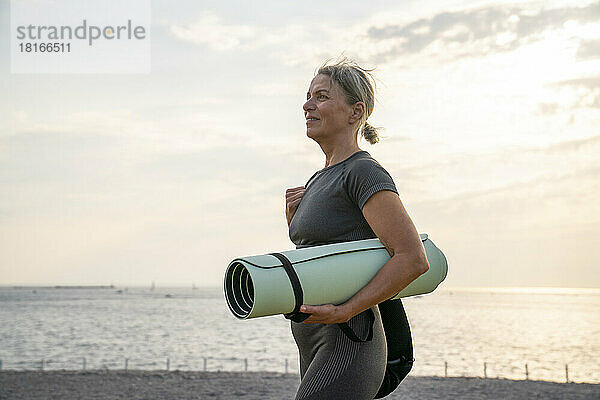 Reife Frau hält bei Sonnenuntergang eine Trainingsmatte in der Hand