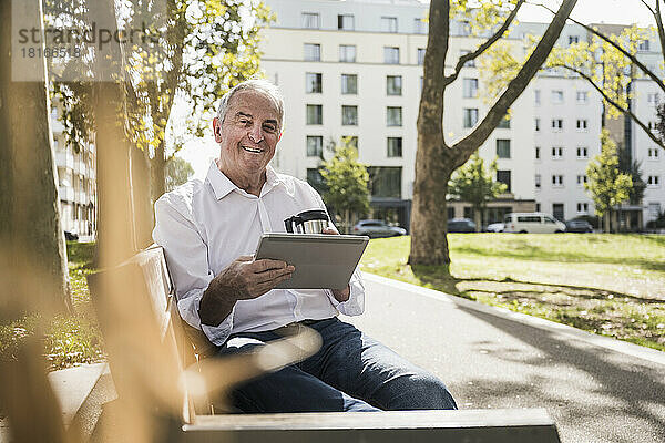 Lächelnder älterer Mann mit Tablet-PC und isoliertem Getränkebehälter sitzt auf der Bank