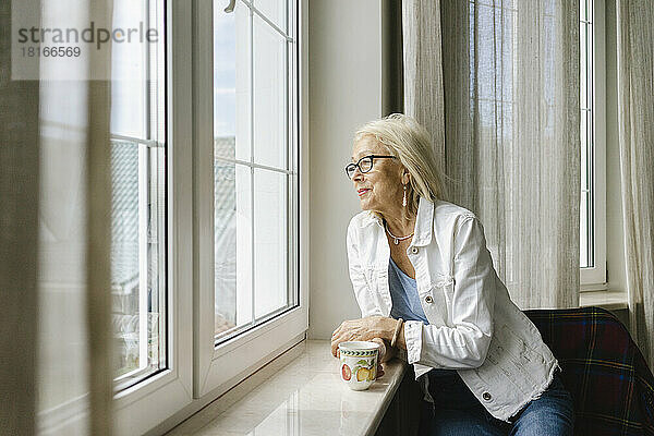 Ältere Frau mit Tasse schaut zu Hause aus dem Fenster