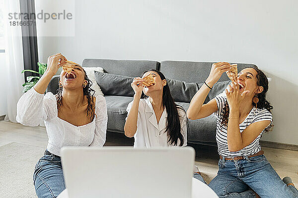 Happy friends eating pizza slices sitting in living room