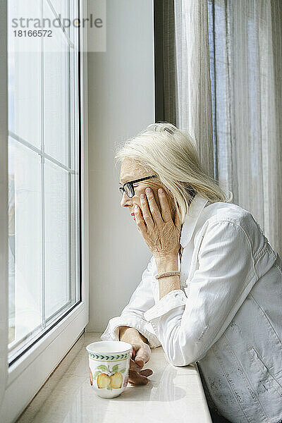Deprimierte ältere Frau mit Tasse lehnt zu Hause auf Fensterbank