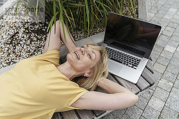 Glückliche blonde Frau mit Laptop auf Bank liegend