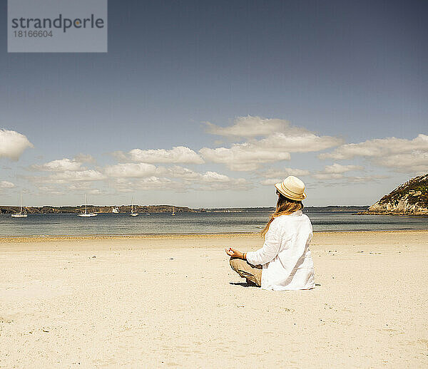 Frau mit Hut praktiziert an einem sonnigen Tag Meditation am Strand