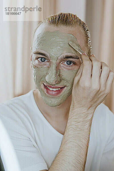 Glücklicher junger Mann  der zu Hause eine Gesichtsmaske aufträgt