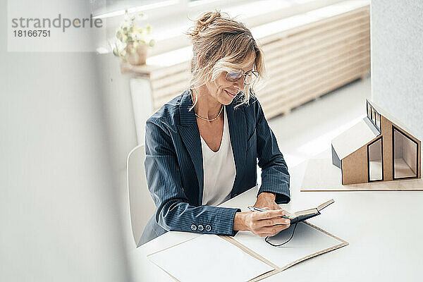 Lächelnde Geschäftsfrau blickt auf das Tagebuch eines Hausmodells auf dem Schreibtisch im Büro