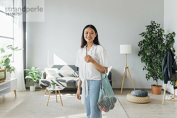 Smiling woman carrying mesh bag with vegetables in living room
