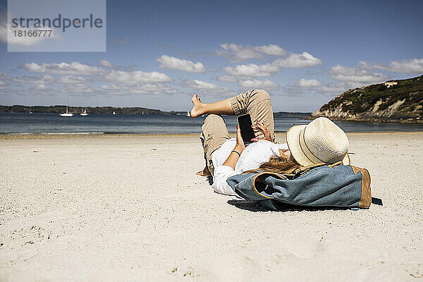 Frau mit Rucksack und Smartphone am Strand liegend