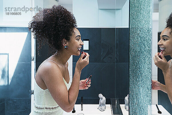Glückliche Frau mit lockigem Haar  die zu Hause im Badezimmer Lippenstift aufträgt