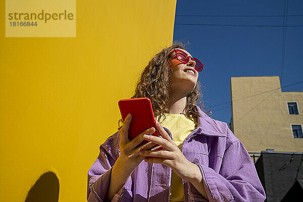 Junge Frau mit Smartphone in der Nähe der Wand an einem sonnigen Tag