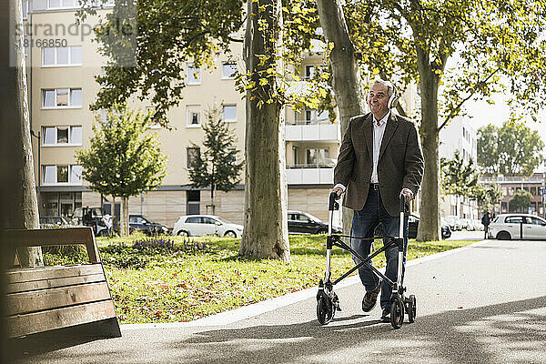 Glücklicher älterer Mann  der mit einem Rollator unterwegs ist und über drahtlose Kopfhörer auf dem Fußweg Musik hört