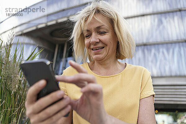 Glückliche reife Frau  die ihr Smartphone vor dem Gebäude benutzt