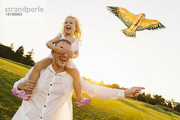 Vater trägt Tochter auf Schultern und lässt bei Sonnenuntergang im Park Drachen steigen