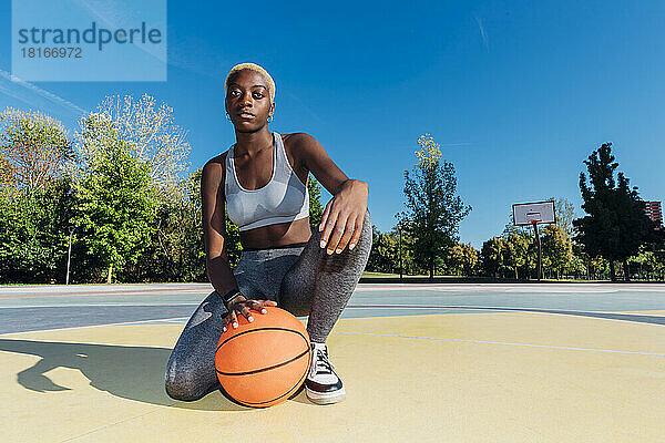 Selbstbewusste Sportlerin kniet mit Ball auf dem Basketballplatz