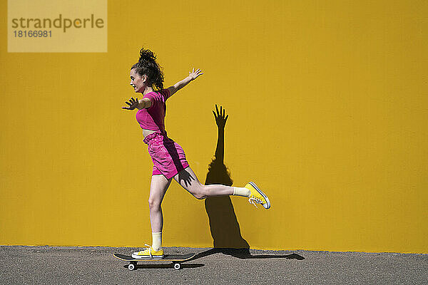 Frau mit ausgestreckten Armen balanciert auf Skateboard vor der Wand