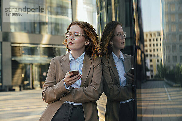 Nachdenkliche Geschäftsfrau mit Smartphone lehnt an Glaswand