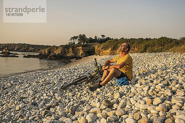 Reifer Mann ruht sich bei Sonnenuntergang mit dem Fahrrad am Ufer aus