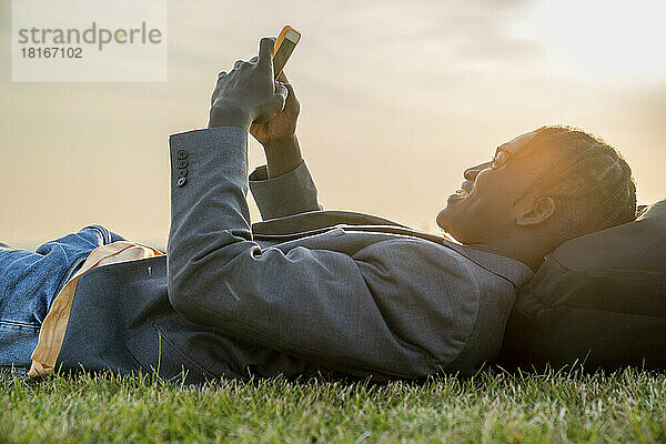 Glücklicher junger Geschäftsmann benutzt Smartphone  das auf Gras liegt