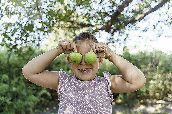 Glückliches Mädchen bedeckt Augen mit grünem Apfel im Park