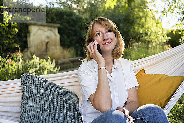 Frau sitzt in Hängematte im Garten und telefoniert mit Mobiltelefon