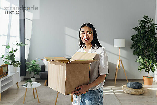 Smiling woman holding cardboard box standing in living room at new home