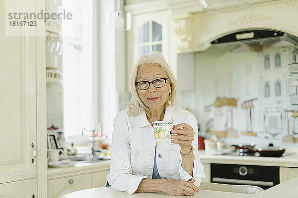Ältere Frau hält eine Tasse und lehnt zu Hause auf der Kücheninsel