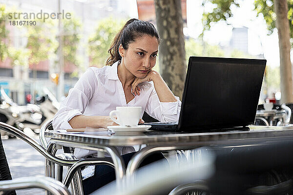 Geschäftsfrau mit der Hand am Kinn arbeitet am Laptop im Straßencafé