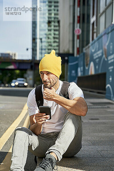 Nachdenklicher Mann mit Smartphone sitzt auf Fußweg