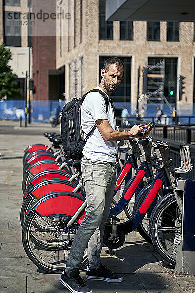 Mann mit Smartphone mietet an sonnigem Tag ein Fahrrad am Parkplatz