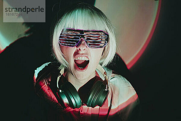 Cheerful woman with neon eyeglasses shouting against black background