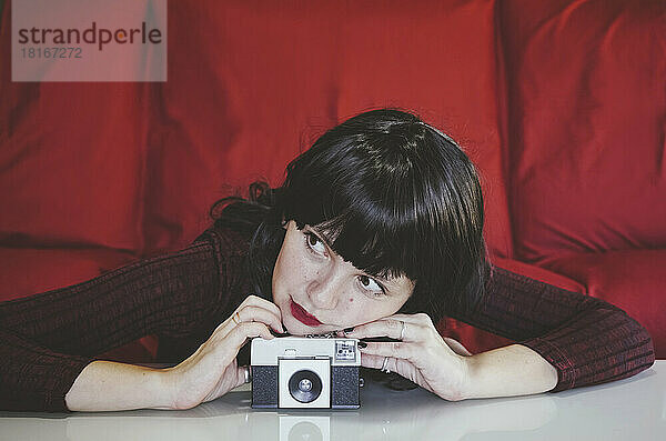 Nachdenkliche Frau lehnt vor einer roten Couch auf eine analoge Kamera