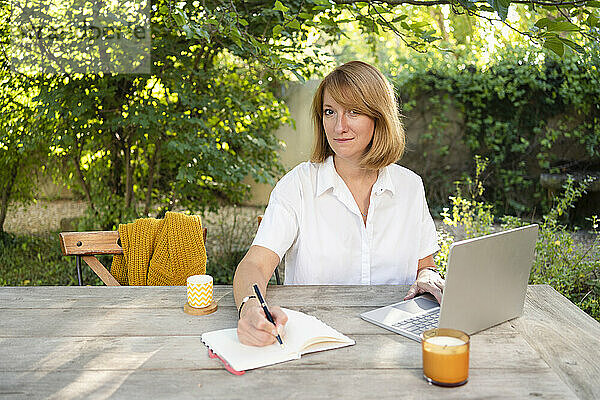 Freiberufler macht sich Notizen  sitzt mit Laptop am Tisch im Garten