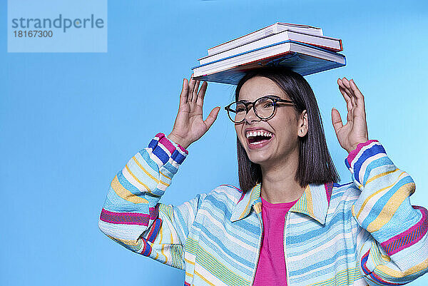 Glückliche junge Frau mit buntem Reißverschluss  die Bücher auf dem Kopf vor blauem Hintergrund balanciert